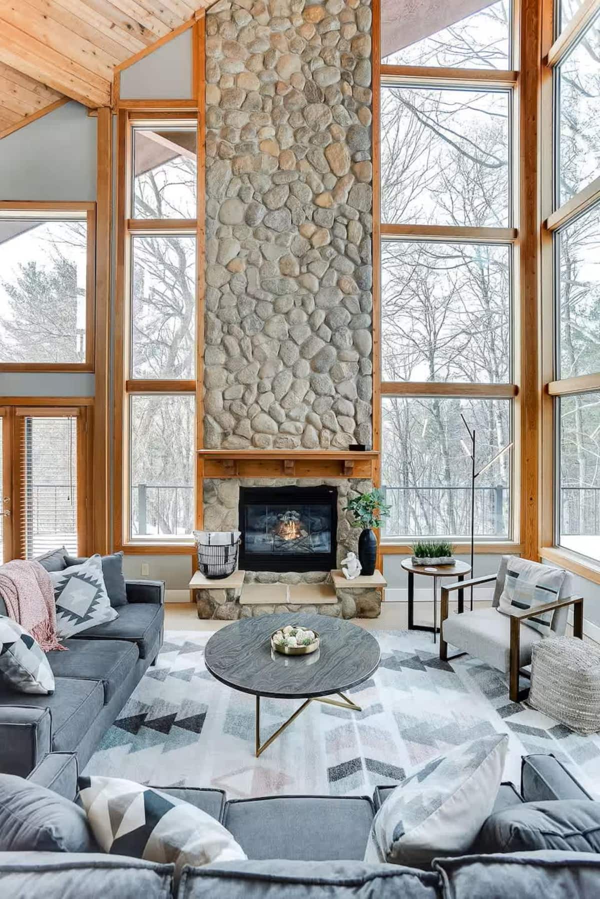 Great room with floor-to-ceiling windows, stone fireplace, and forest views